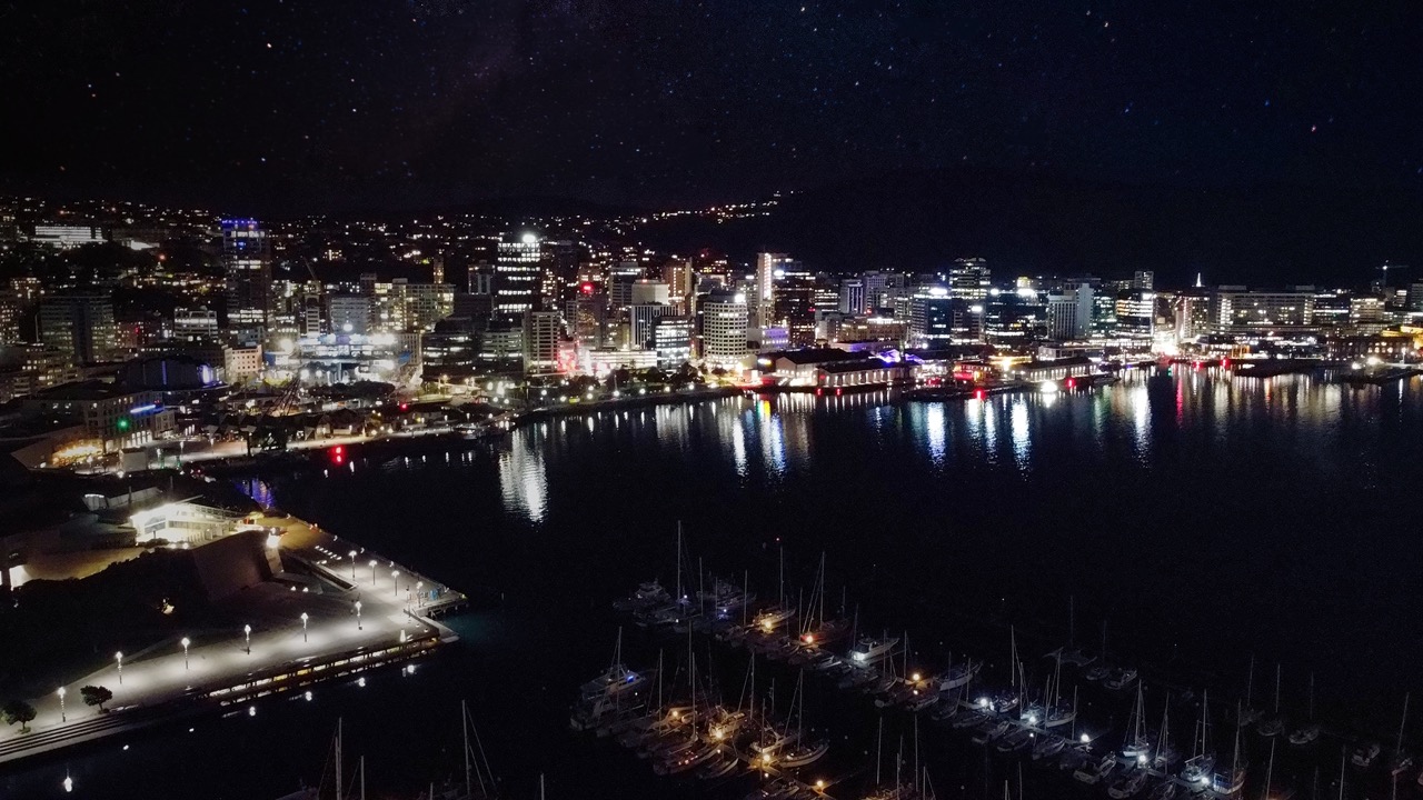 Drone photo of Wellington waterfront at night, featuring illuminated buildings, harbour reflections, and city lights.